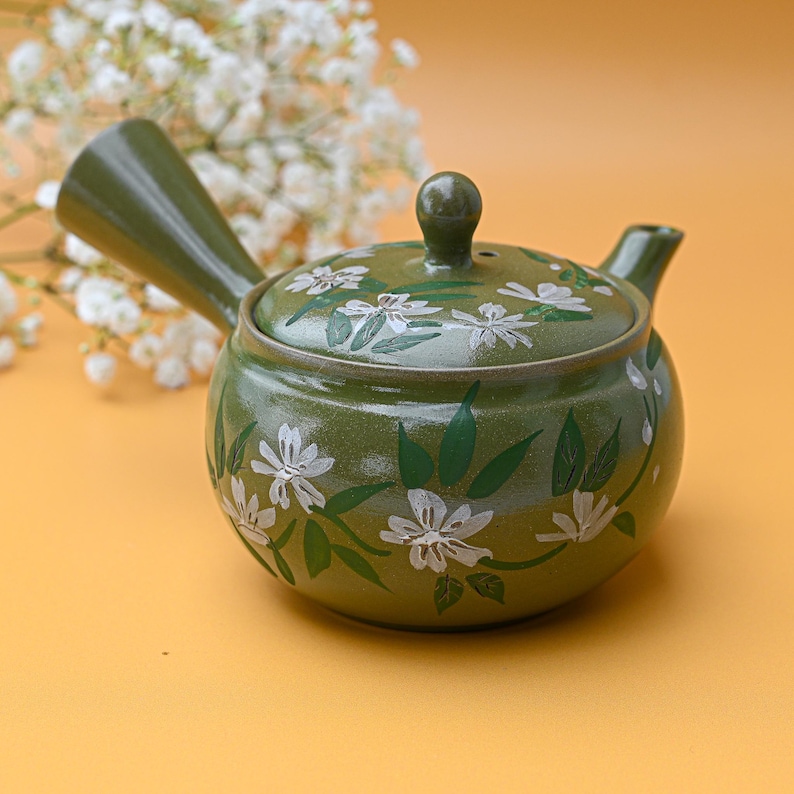 Puede incluir: Una tetera de cer&aacute;mica verde con un dise&ntilde;o floral blanco. La tetera tiene un pico y una asa.