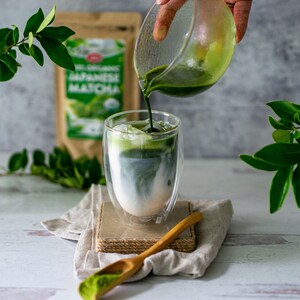 May include: A glass of matcha latte with ice, being poured from a glass pitcher. A wooden spoon with matcha powder sits on a linen cloth. A bag of Japanese matcha is in the background.