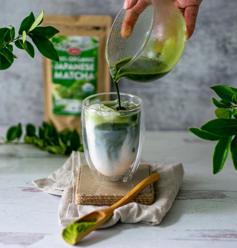 May include: A glass of matcha latte with ice, being poured from a glass pitcher. A wooden spoon with matcha powder sits on a linen cloth. A bag of Japanese matcha is in the background.