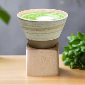 May include: A ceramic cup with a light brown and white striped design, filled with a green matcha latte, sits on a square, light brown ceramic base.