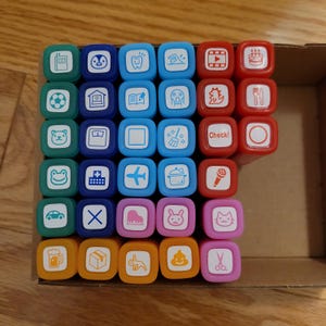 May include: A set of 48 colorful rubber stamps with various images, including a soccer ball, a house, a book, a ghost, a dinosaur, a fork and knife, a frog, a plus sign, an airplane, a pot, a microphone, a car, an X, a piano, a bunny, a cat, a beer mug, a box, a unicorn, and poop. The stamps are arranged in a grid pattern in a wooden box.
