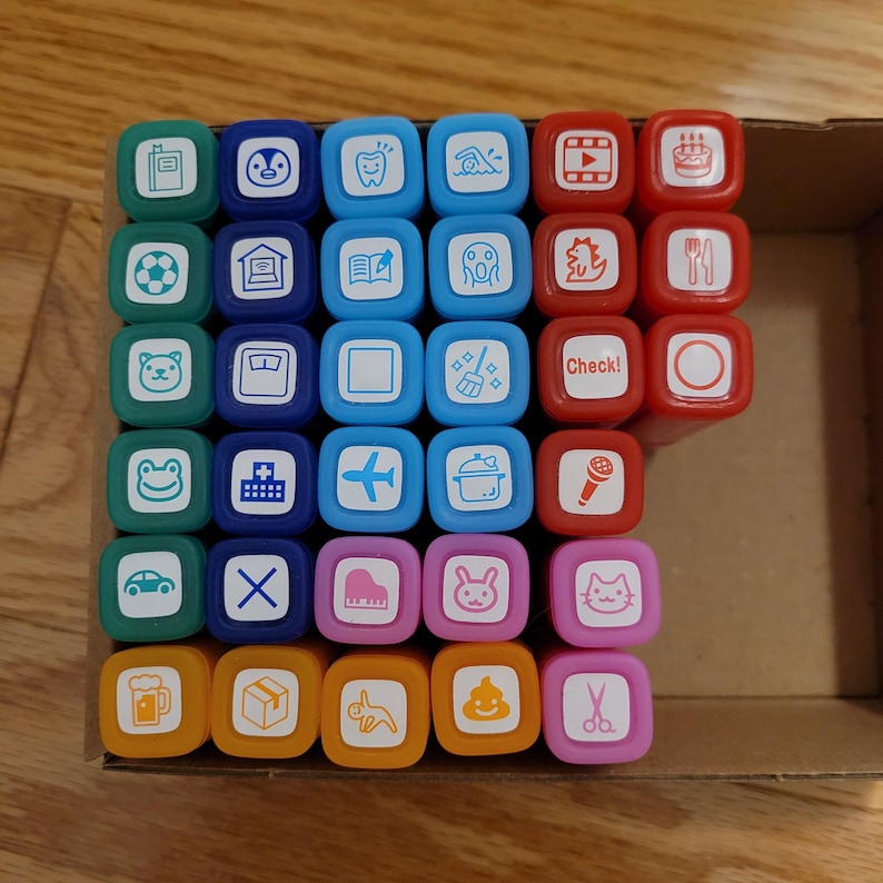 May include: A set of 48 colorful rubber stamps with various images, including a soccer ball, a house, a book, a ghost, a dinosaur, a fork and knife, a frog, a plus sign, an airplane, a pot, a microphone, a car, an X, a piano, a bunny, a cat, a beer mug, a box, a unicorn, and poop. The stamps are arranged in a grid pattern in a wooden box.