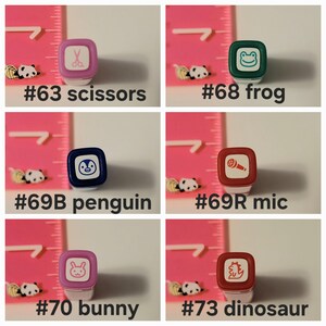 May include: Six colorful rubber stamps with different designs. The stamps are arranged in two rows of three. The top row includes a pair of scissors, a frog, and a microphone. The bottom row includes a penguin, a bunny, and a dinosaur.