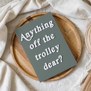 May include: A rectangular greeting card with a solid sage green background and white text that reads Anything off the trolley dear? The card is positioned on a round wooden tray, which is set against a backdrop of white fabric.