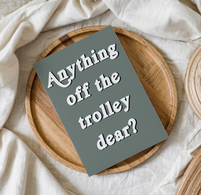 May include: A rectangular greeting card with a solid sage green background and white text that reads Anything off the trolley dear? The card is positioned on a round wooden tray, which is set against a backdrop of white fabric.