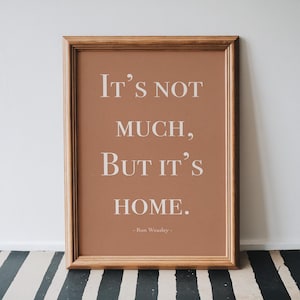 May include: A rectangular wooden frame containing a terracotta-coloured print with the white text "IT'S NOT MUCH, BUT IT'S HOME." followed by "- Ron Weasley -" in a smaller font size, centred within the frame.