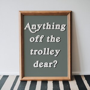 May include: A framed wall art print featuring a dark sage green background with the white text "Anything off the trolley dear?" in a bold, rounded font. The frame is made of light brown wood with a simple, clean profile and a visible grain texture.