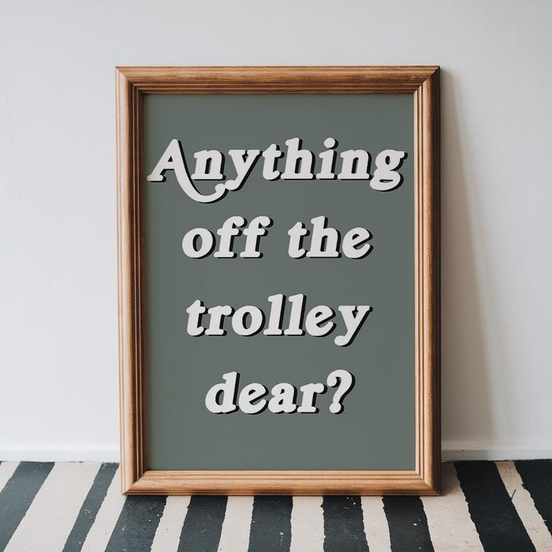 May include: A framed wall art print featuring a dark sage green background with the white text "Anything off the trolley dear?" in a bold, rounded font. The frame is made of light brown wood with a simple, clean profile and a visible grain texture.
