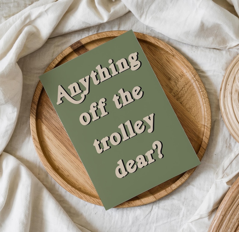 May include: A rectangular greeting card with a sage green background and the text "Anything off the trolley dear?" printed in a bold, cream-colored font with a dark shadow effect, positioned diagonally across the center of the card.
