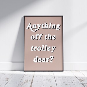 May include: A rectangular framed wall art print featuring a dusty rose background with the white text Anything off the trolley dear? written in a bold, rounded, sans-serif font. The frame is thin and black, surrounding the centered text design.