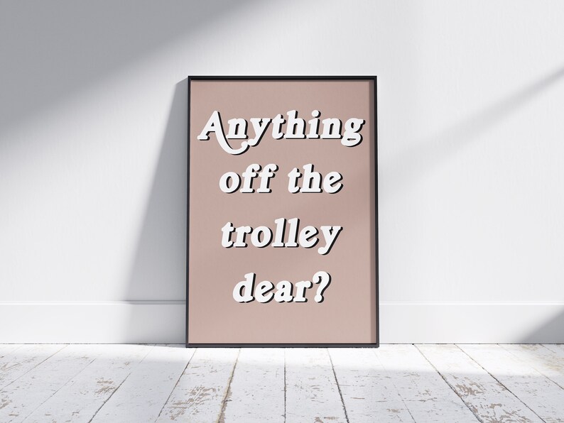 May include: A rectangular framed wall art print featuring a dusty rose background with the white text Anything off the trolley dear? written in a bold, rounded, sans-serif font. The frame is thin and black, surrounding the centered text design.