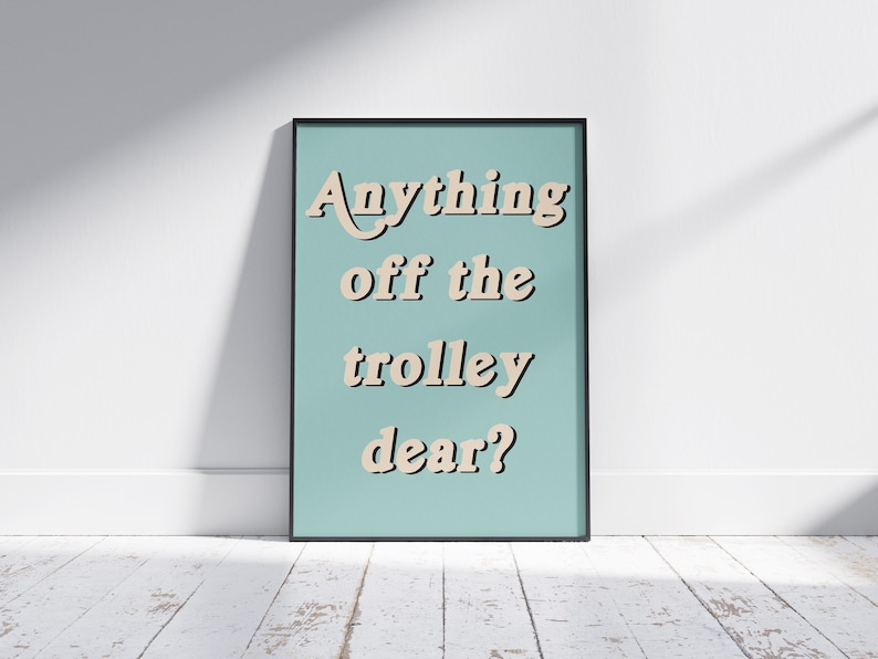 May include: A framed wall art print featuring a light blue background with the text Anything off the trolley dear? written in a beige, bold, sans-serif font with a dark drop shadow effect. The frame is thin and black, surrounding the rectangular print.