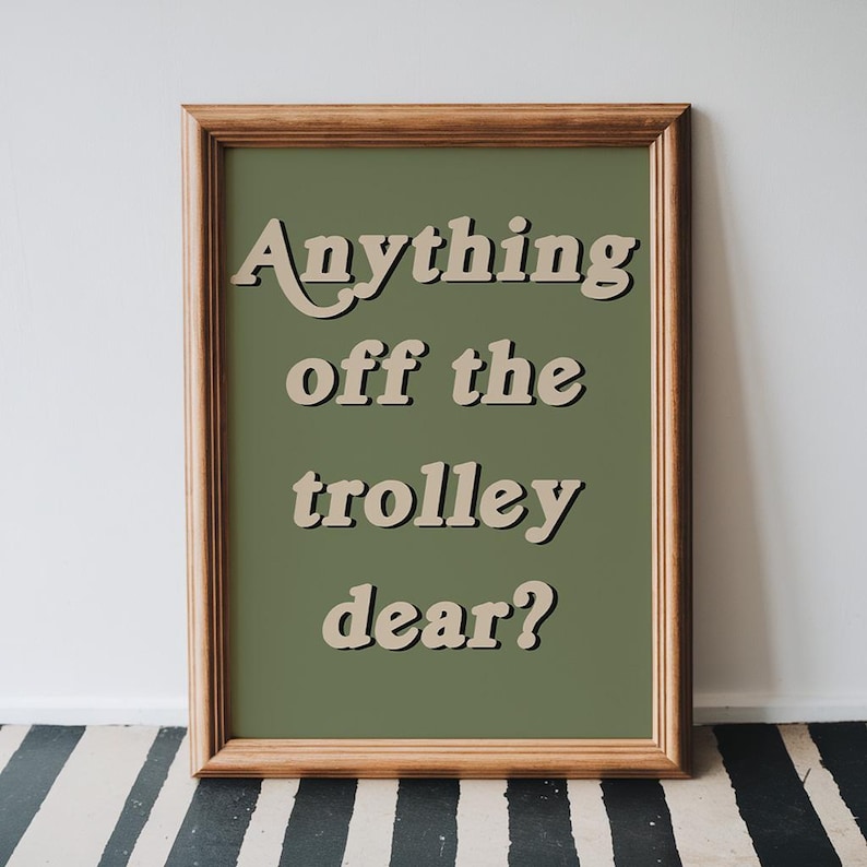 May include: A framed wall art print featuring a solid olive green background with the beige text "Anything off the trolley dear?" in a bold, retro-inspired font. The print is enclosed in a simple, light-toned wooden frame with a classic profile.
