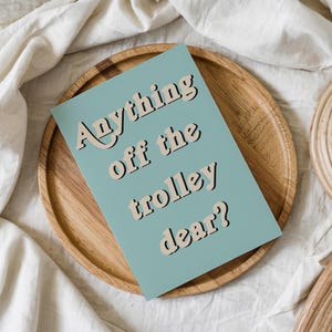 May include: A rectangular card with a muted blue background featuring the text Anything off the trolley dear? in a bold, beige, retro-style font with a dark outline. The card is positioned centrally on a round, light-colored wooden tray.