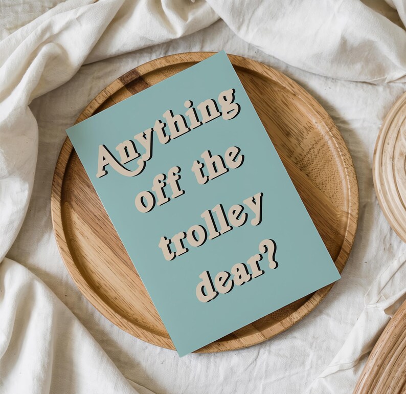 May include: A rectangular card with a muted blue background featuring the text Anything off the trolley dear? in a bold, beige, retro-style font with a dark outline. The card is positioned centrally on a round, light-colored wooden tray.