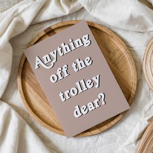 May include: A rectangular greeting card with a muted mauve background featuring the white text Anything off the trolley dear? in a bold, sans-serif font with a subtle black drop shadow, positioned centrally on the card surface.