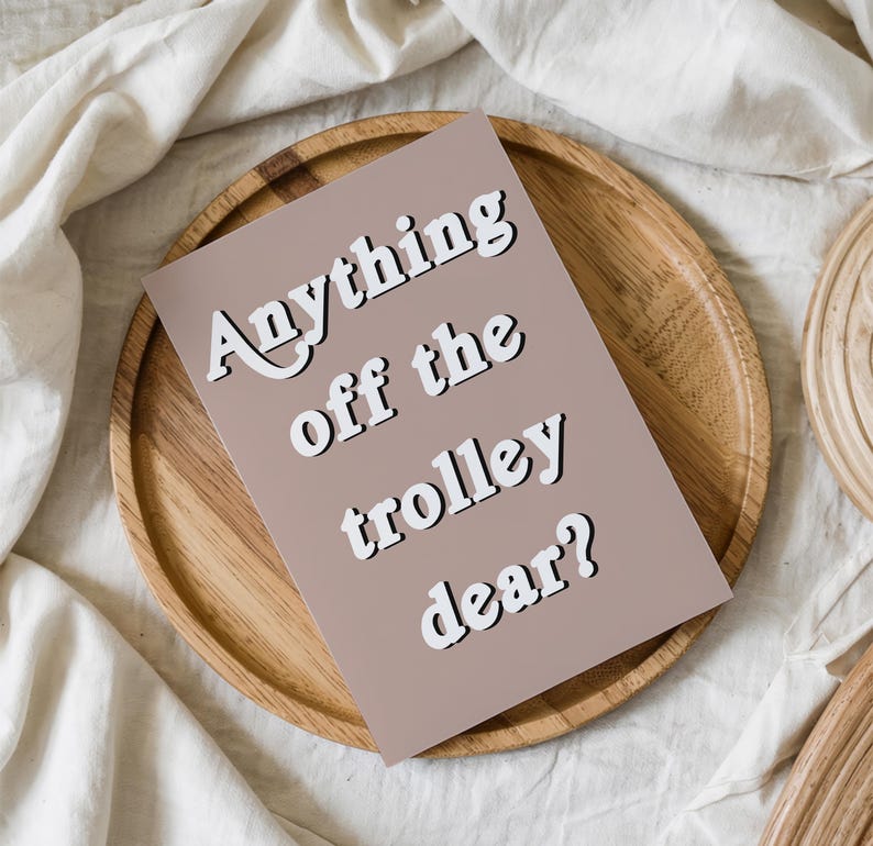 May include: A rectangular greeting card with a muted mauve background featuring the white text Anything off the trolley dear? in a bold, sans-serif font with a subtle black drop shadow, positioned centrally on the card surface.