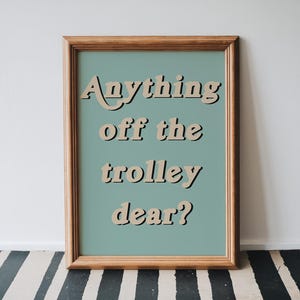 May include: A framed wall art print featuring a muted sage green background with the text Anything off the trolley dear? printed in a beige, bold, retro-style font with a dark drop shadow, enclosed in a simple, light-toned wooden frame.