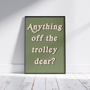 May include: A rectangular framed wall art piece featuring a solid olive green background with the text "Anything off the trolley dear?" printed in a cream-colored, bold, serif font with a subtle drop shadow effect, enclosed in a thin black frame.