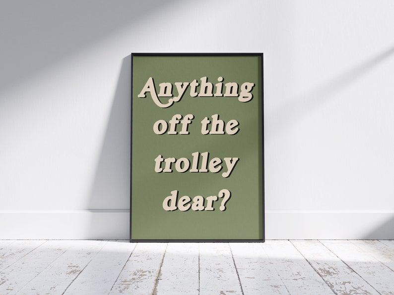 May include: A rectangular framed wall art piece featuring a solid olive green background with the text "Anything off the trolley dear?" printed in a cream-colored, bold, serif font with a subtle drop shadow effect, enclosed in a thin black frame.