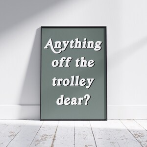 May include: A rectangular wall art print in a thin black frame featuring a muted sage green background with the white text Anything off the trolley dear? displayed in a bold, serif font. The lettering is centered and stacked in four lines.