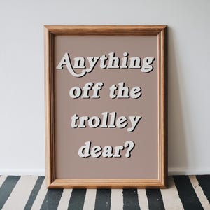 May include: A rectangular wooden frame containing a print with a muted mauve background. The text "Anything off the trolley dear?" is written in a white, bold, sans-serif font with a black drop shadow, centered within the frame.