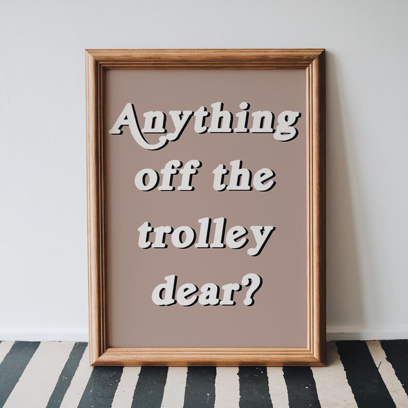 May include: A rectangular wooden frame containing a print with a muted mauve background. The text "Anything off the trolley dear?" is written in a white, bold, sans-serif font with a black drop shadow, centered within the frame.