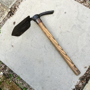 May include: A mattock tool featuring a dark metal head with a pointed pick on one side and a flat blade on the other, securely attached to a long, straight, light brown wooden handle with a visible grain pattern and a metallic collar.