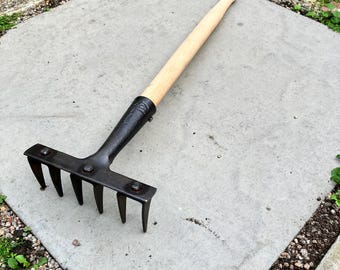 May include: A hand rake featuring a long, light-colored wooden handle attached to a black metal head with five sturdy, downward-curving tines. The metal head is secured to the handle with a metal collar and visible rivets for stability.