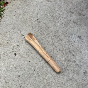 Può includere: Un oggetto di legno di colore chiaro, forse un manico di attrezzo o un pezzo decorativo, è appoggiato su una superficie di cemento. L'oggetto ha una forma cilindrica con una parte superiore più ampia e un fondo più stretto e arrotondato. È fissato con sottili fasce scure.