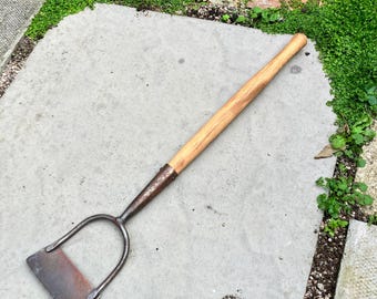 May include: A garden hoe featuring a long, cylindrical wooden handle attached to a dark, weathered metal head. The metal component consists of a U-shaped frame connected to a flat, rectangular blade with visible signs of oxidation.