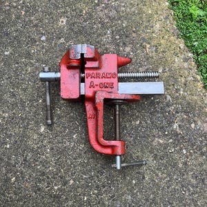 May include: A red PARAMO A-ONE bench vise, A1 model, with a silver screw and handle. The vise is designed to clamp and hold objects securely. The tool is shown on a textured gray surface.