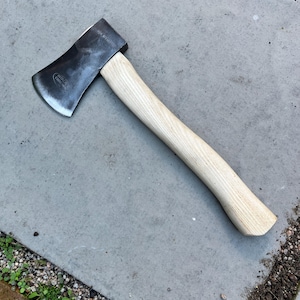 May include: A hatchet with a dark grey metal head and a light-coloured wooden handle. The metal head features a curved blade edge and a visible stamped logo. The handle is smooth, curved, and shows a natural wood grain pattern throughout its length.