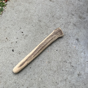Può includere: Un bastone di legno di colore chiaro, forse un manico di attrezzo, giace su una superficie di cemento. Il legno presenta un motivo a venature naturali con striature marroni più scure. L'oggetto è cilindrico con un'estremità arrotondata.
