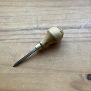May include: A woodworking awl with a light-colored wooden handle and a dark metal blade. The handle has a rounded shape with carved details and a brass ferrule. The awl is designed for making holes in wood or other materials.