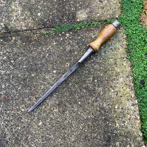 May include: A vintage metal chisel with a wooden handle. The chisel has a long, tapered blade and a rounded wooden handle with a metal ferrule. The handle is a light brown color, and the blade is a silver color. The chisel is lying on a concrete surface.