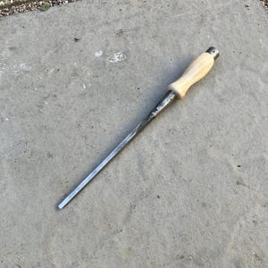 May include: A metal chisel with a wooden handle. The chisel has a long, flat, silver blade and a light-colored wooden handle. The tool is designed for woodworking or similar tasks. The chisel is lying on a concrete surface.