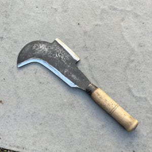 Peut inclure: Un outil serpette doté d'une lame métallique sombre et incurvée avec un tranchant affûté et un manche droit en bois clair. La surface métallique présente des signes d'usure et de texture, tandis que le manche est de forme cylindrique.