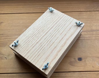 May include: A wooden flower press with four wing nuts. The press is made of light-colored wood and is rectangular. The wing nuts are silver and are located at each corner of the press. The press is sitting on a wooden surface.