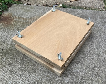 May include: A rectangular flower press constructed from two light-colored wooden boards. The boards are held together by four metal bolts positioned at each corner, each secured with a silver-toned metal wing nut for tightening the press.
