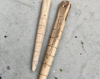 May include: Two tapered wooden tools with rounded ends and engraved horizontal lines along their length. The left tool is light-colored and smooth, while the right tool is darker with visible wood grain and a small hole near the top end.