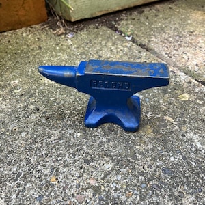 May include: A small, blue anvil with the word "RECORD" stamped on the side. The anvil is made of metal and has a classic shape, with a pointed horn on one end and a flat surface on top. It sits on a concrete surface.