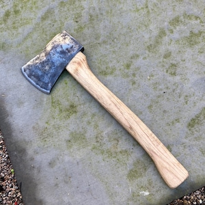 May include: A weathered axe with a wooden handle and a steel blade. The axe head is a dark gray color, and the wooden handle is a light brown. The axe is lying on a gray stone surface.