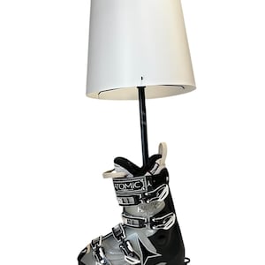 May include: A white lampshade with a black metal pole attached to a pair of black and silver ski boots. The boots have the brand name "ATOMIC" printed on them.
