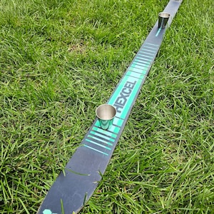 Op de afbeelding: Een zwarte ski met een groen-wit ontwerp en het woord "EXCEL" erop gedrukt. Vier kleine zilveren bekers zijn op de ski geplaatst.