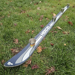 Op de afbeelding: Een blauwe en zilveren ski met het woord "HEXCEL" erop gedrukt. Vier kleine zilveren bekers zijn op de ski geplaatst, gelijkmatig verdeeld.