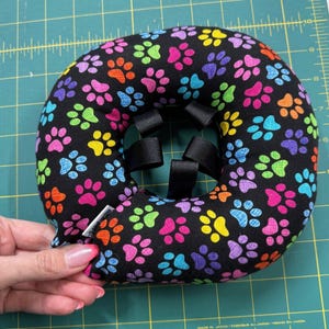 May include: A black pet recovery collar with a colorful paw print pattern. The collar is padded and has adjustable black straps. The paw prints are in shades of pink, orange, yellow, green, blue, and purple.