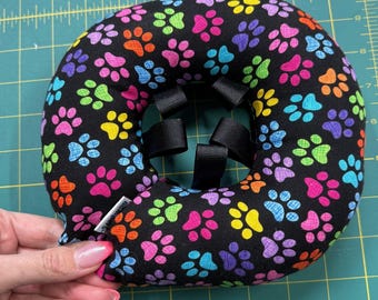 May include: A black pet recovery collar with a colorful paw print pattern. The collar is padded and has adjustable black straps. The paw prints are in shades of pink, orange, yellow, green, blue, and purple.