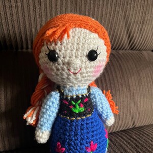 May include: A crocheted doll with orange hair, white skin, and blue eyes. The doll is wearing a blue dress with a black and green vest and pink flowers.