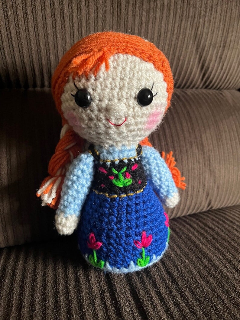 May include: A crocheted doll with orange hair, white skin, and blue eyes. The doll is wearing a blue dress with a black and green vest and pink flowers.
